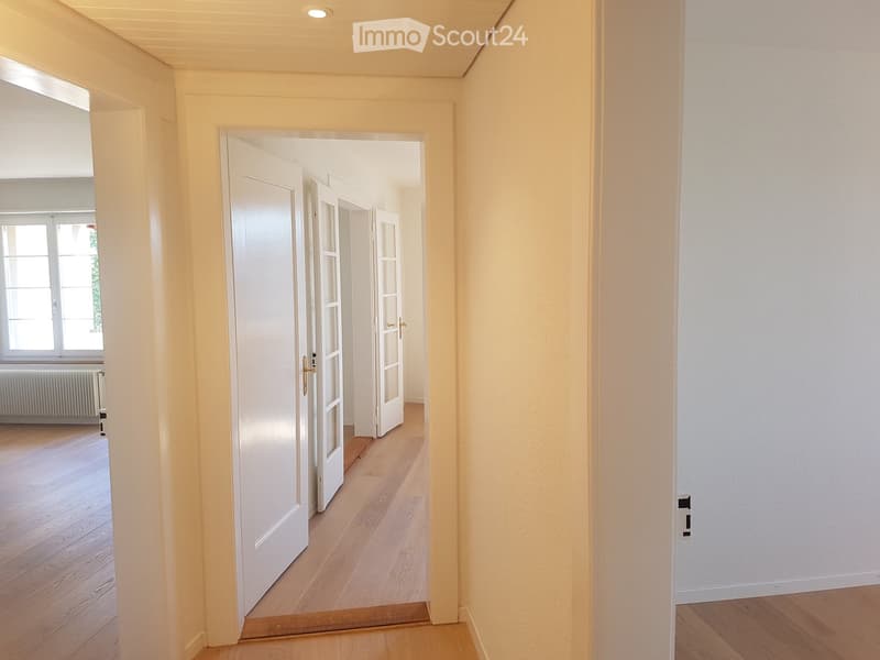 Empty hallway with wooden flooring, white walls, and open doors leading to another room.