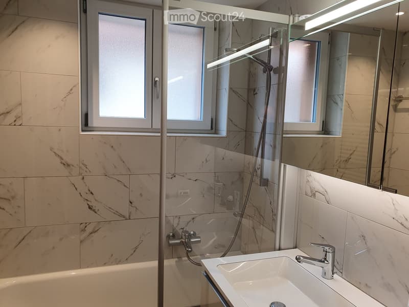 Modern bathroom with marble tiles, a bathtub, a sink with a chrome faucet, a large mirror, and a glass-enclosed shower area with a modern showerhead.