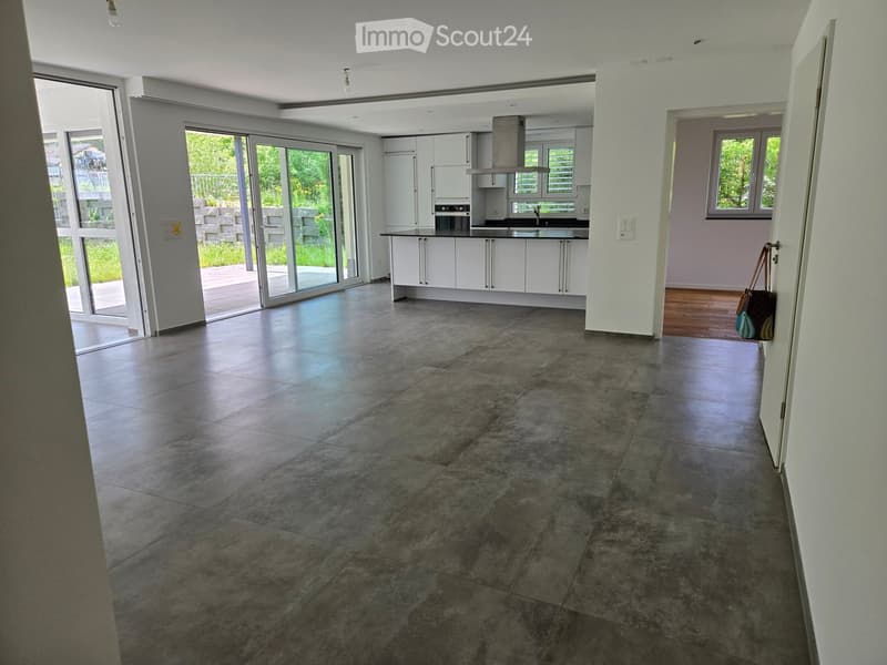 Open-plan space with a modern kitchen area featuring white cabinets, a central island, and large sliding glass doors leading to an outdoor area. The floor is made of large, dark grey tiles.