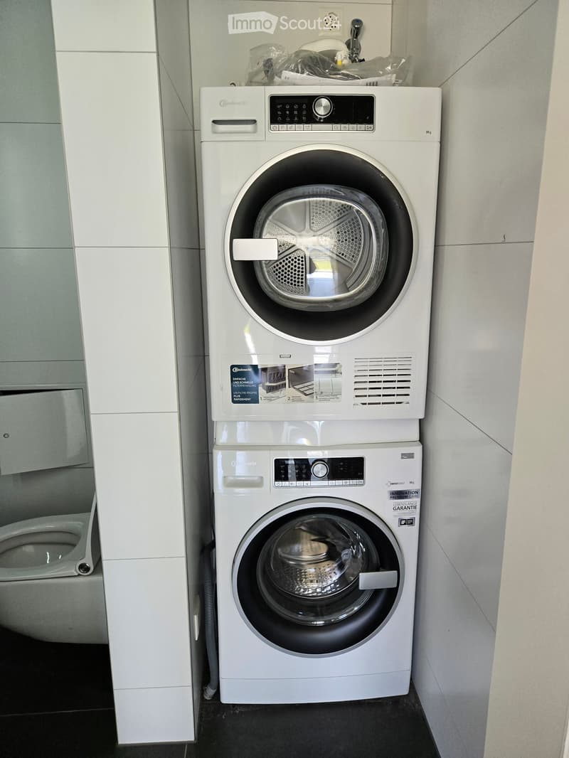 Modern bathroom with a white washing machine and dryer stacked vertically, a toilet, and sleek white cabinetry.