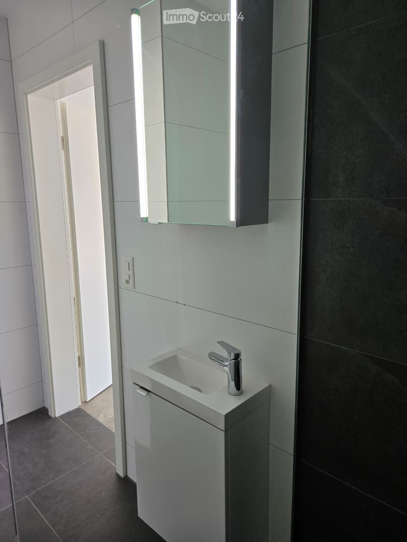 Modern bathroom with a white sink, sleek faucet, and illuminated mirror above it. Dark tiled walls and floor.