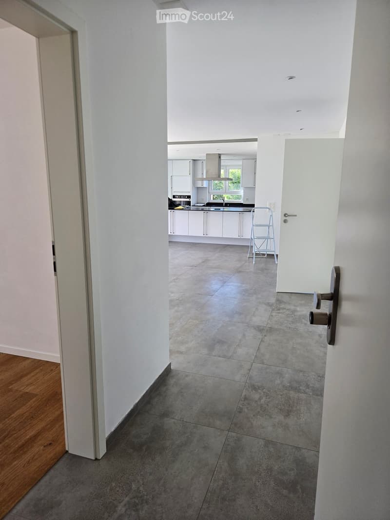 Empty hallway with grey tiled floor, white walls, and wooden flooring on one side. A doorway leads to a modern kitchen with white cabinets and stainless steel appliances.