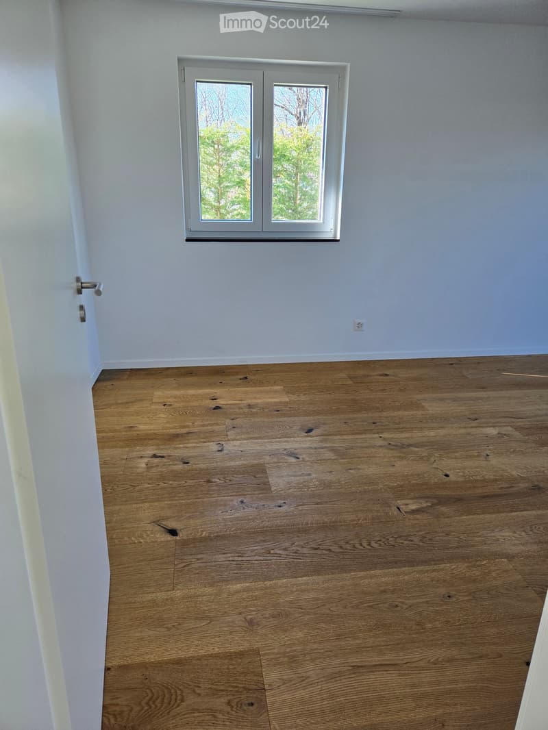 Empty room with light wooden flooring and white walls, featuring a large window with a view of trees and a door with a silver handle.