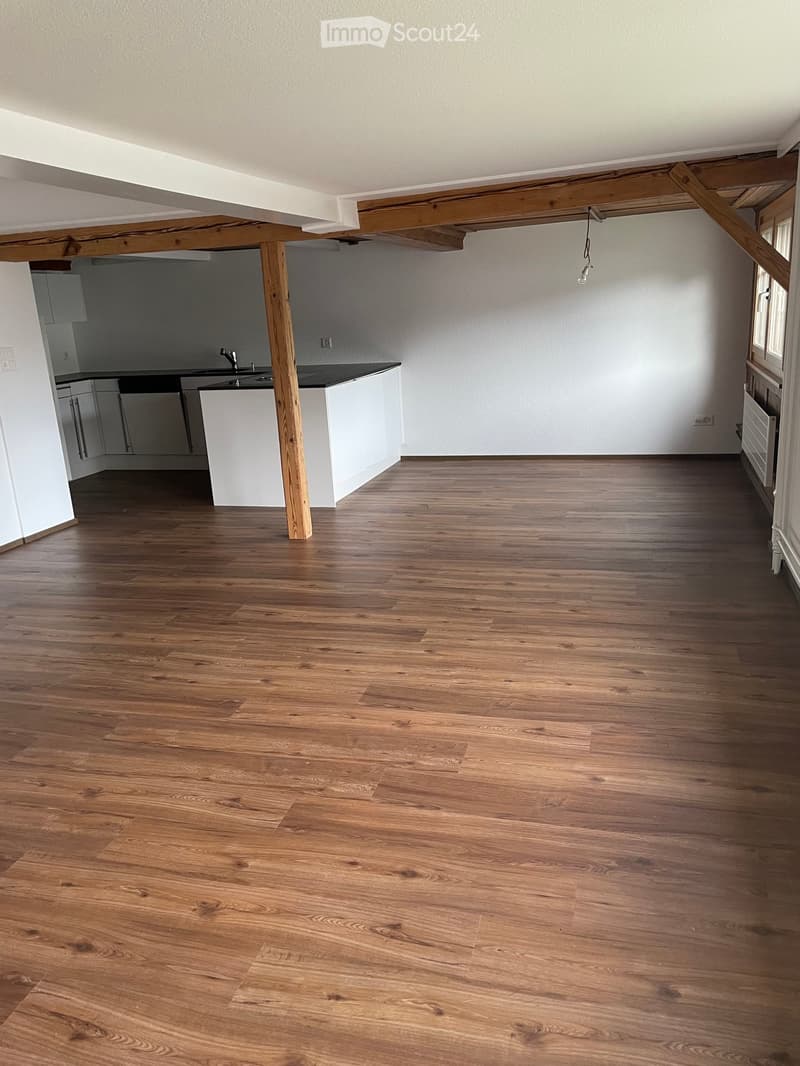 Empty room with wooden flooring, white walls, and a small kitchen area visible in the background.