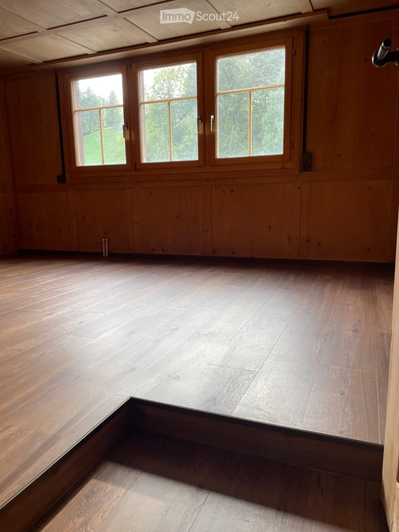 Empty room with wooden walls, wooden floor, large windows, and exposed wooden ceiling beams.