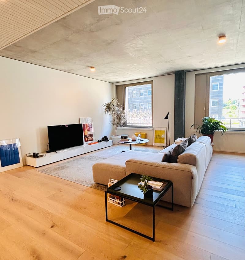 Modern living room with light wooden flooring, a large sectional sofa, a black coffee table, a flat-screen TV on a white stand, and large windows offering a city view.