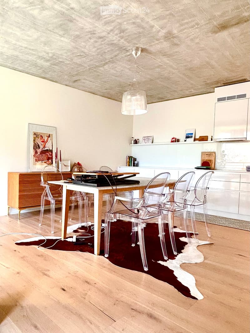 Modern kitchen with a white dining table, four transparent chairs, a wooden cabinet, and a hanging lamp. The floor is light wood, and a red and white cowhide rug is placed under the table.