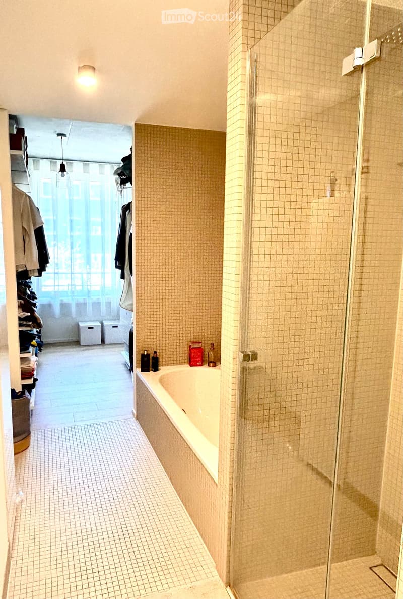 Bathroom with a bathtub, glass-enclosed shower, tiled walls, and a small storage area with clothing and bins.