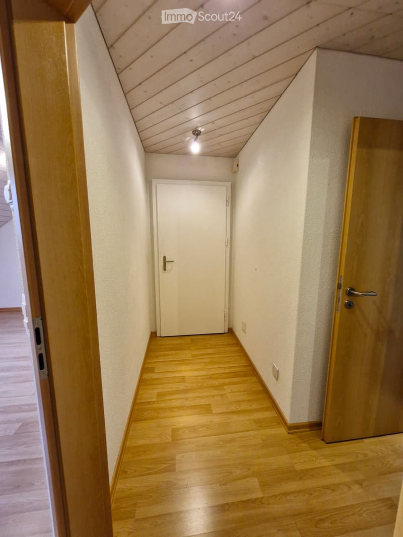A narrow hallway with wooden flooring, white walls, and a single closed door at the end. The ceiling has exposed wooden beams and a single light fixture.