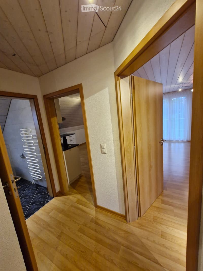 A narrow hallway with wooden flooring and white walls, featuring several open doors leading to different rooms, including a bathroom and a kitchen area.