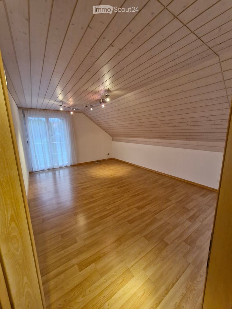 Empty room with wooden flooring and slanted wooden ceiling, featuring recessed lighting and a window with white curtains.