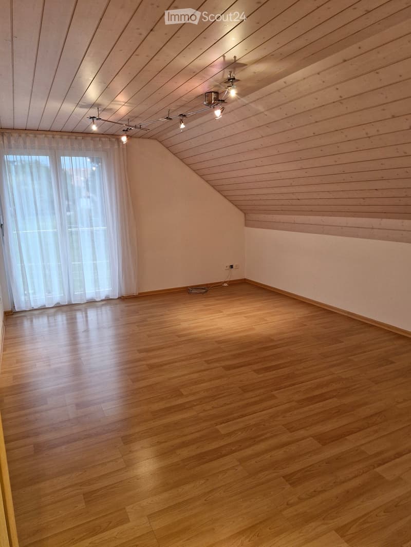 Empty room with wooden flooring, white walls, and a slanted wooden ceiling. Features include a window with white curtains and recessed lighting fixtures.