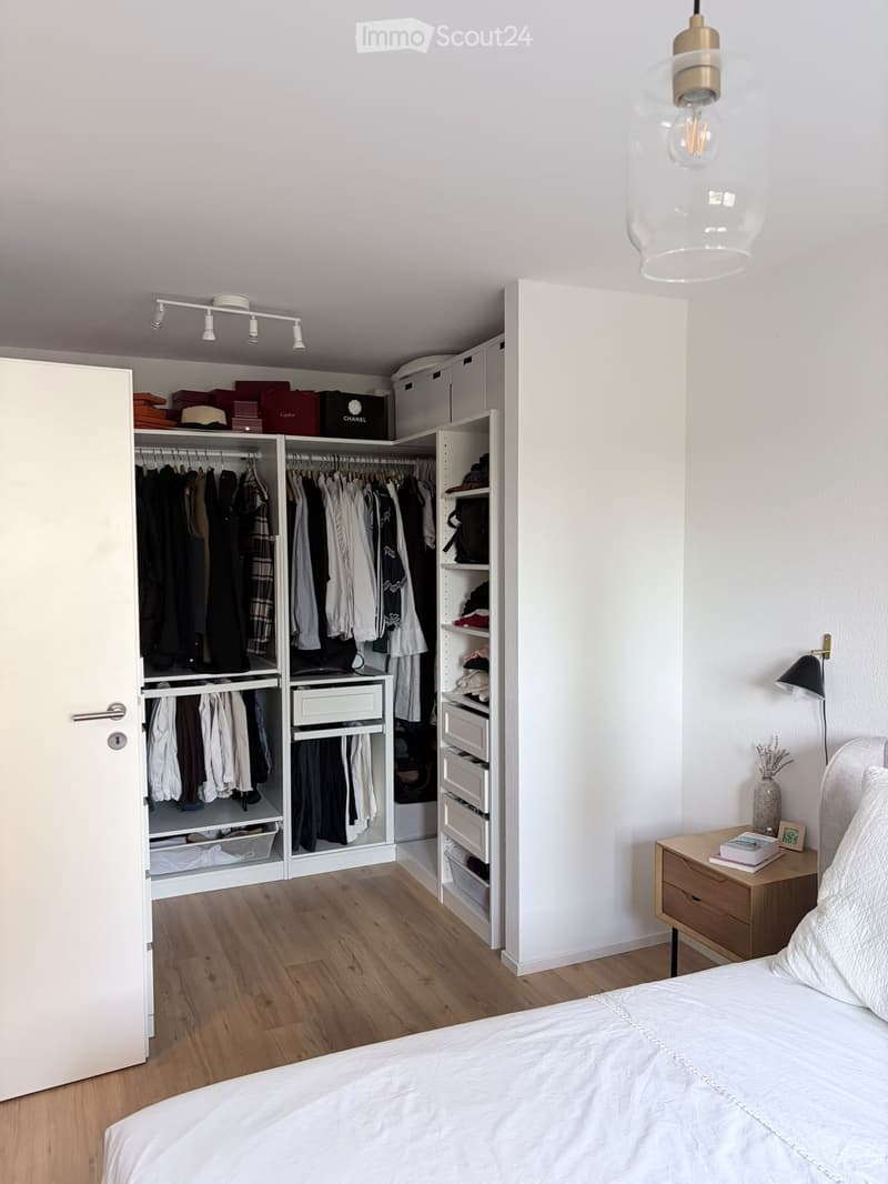 Modern bedroom with a large bed, nightstand, wardrobe with hanging clothes, and a pendant light fixture.