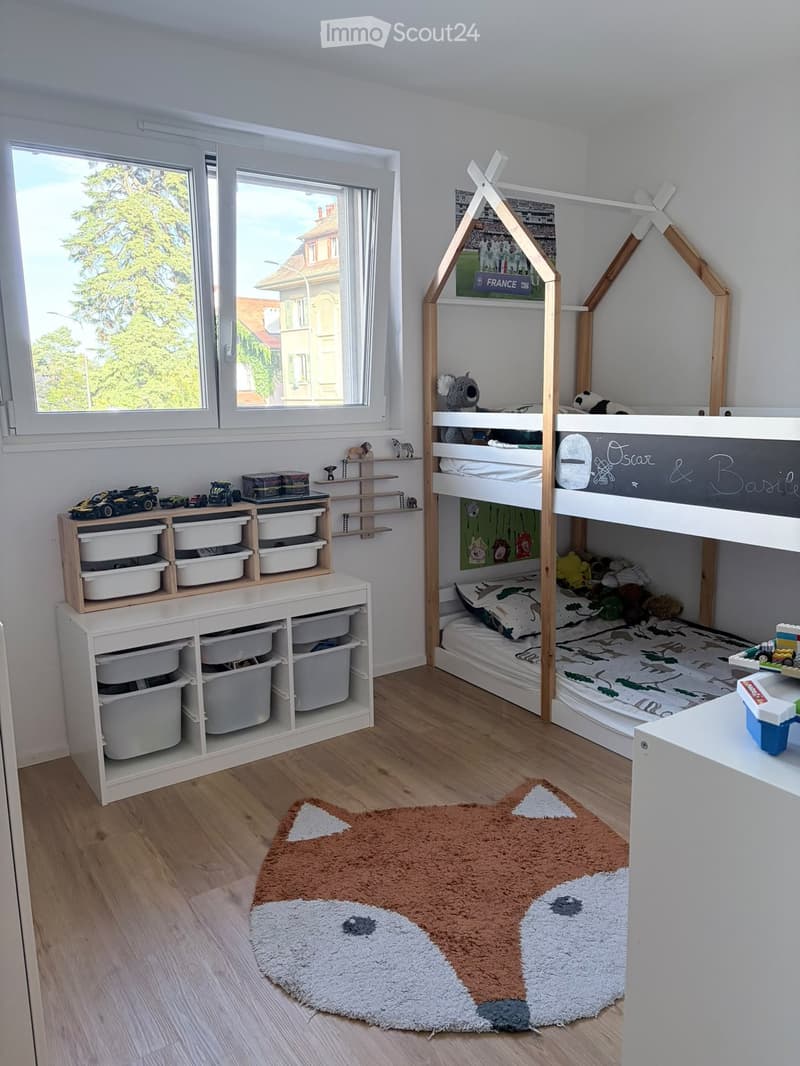 Bright, modern children's bedroom with a bunk bed, storage shelves, toys, and a large window overlooking a tree-lined street.