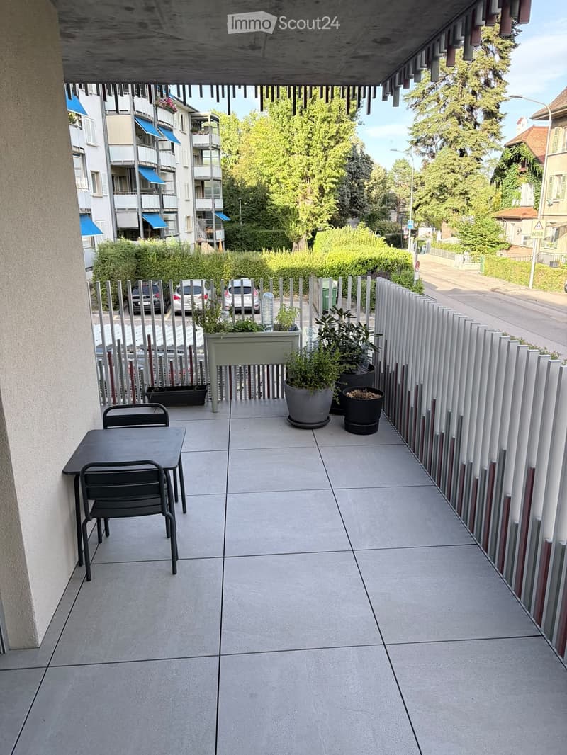 Balcony with tiled floor, metal table and chairs, potted plants, and a view of the street and surrounding buildings.
