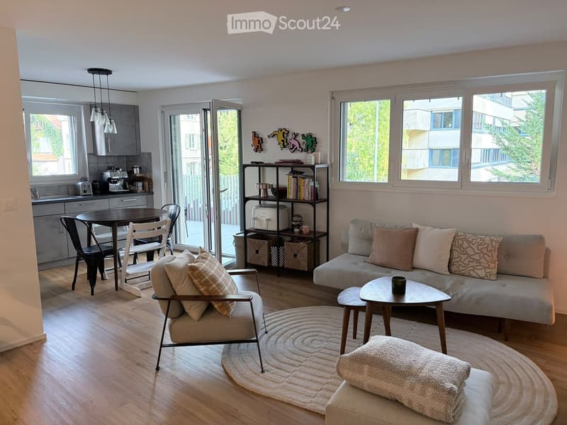 A modern, open-concept living and dining area with a sofa, coffee table, armchair, dining table, and kitchen area. The space features large windows, wooden flooring, and decorative pillows on the sofa.