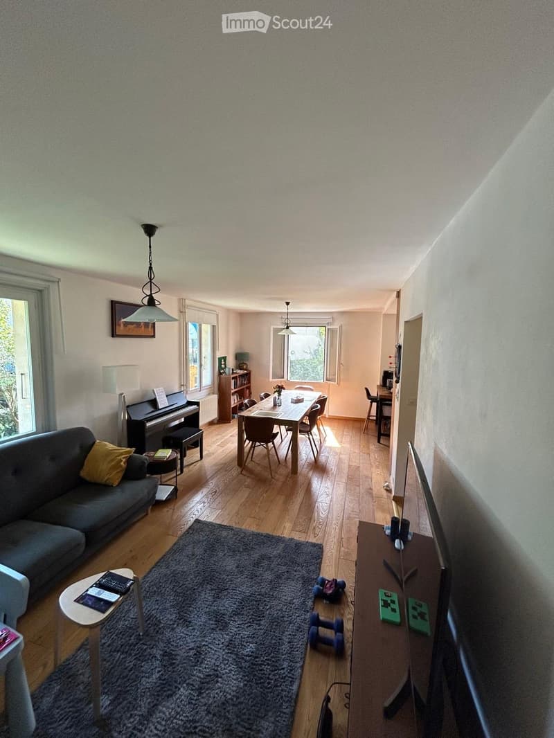 A spacious living room with wooden flooring, a grey sofa with yellow cushions, a coffee table, a large rug, a dining table with chairs, a piano, a bookshelf, and a TV stand with a screen.