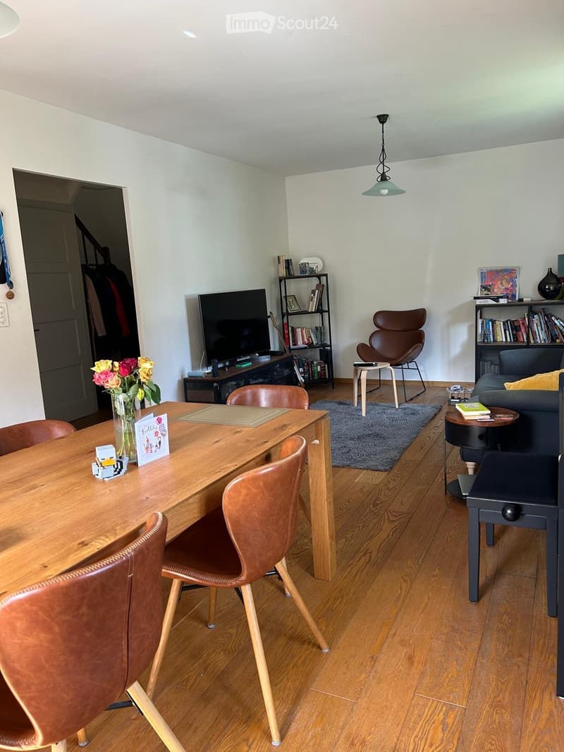 A well-furnished room with a wooden dining table, leather chairs, a sofa, a television, bookshelves, and a small side table with a vase of flowers.
