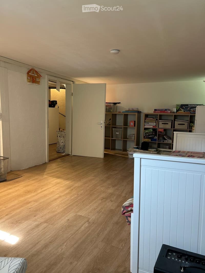 A small, well-lit room with wooden flooring and white walls. A shelf with books and storage boxes is visible, along with a small table and a partially visible bed with a patterned blanket.