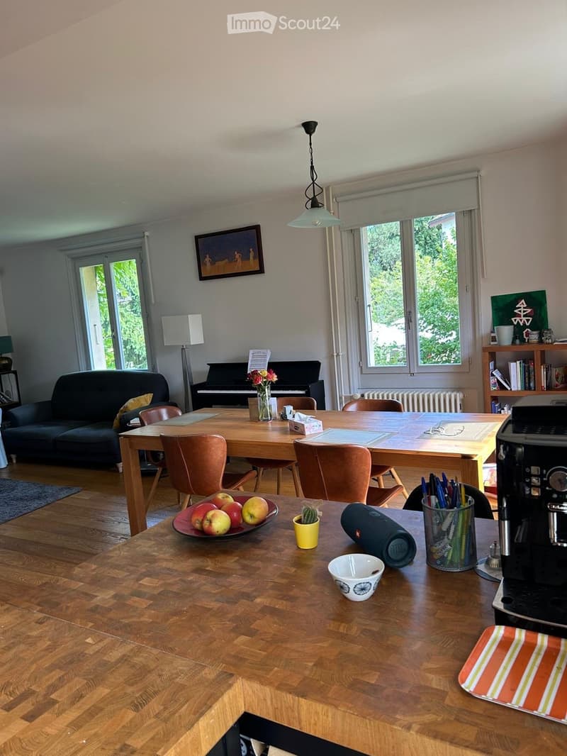 Bright living room with wooden flooring, large windows, a sofa, a dining table with chairs, a piano, and a coffee area with a coffee maker and various items on the counter.