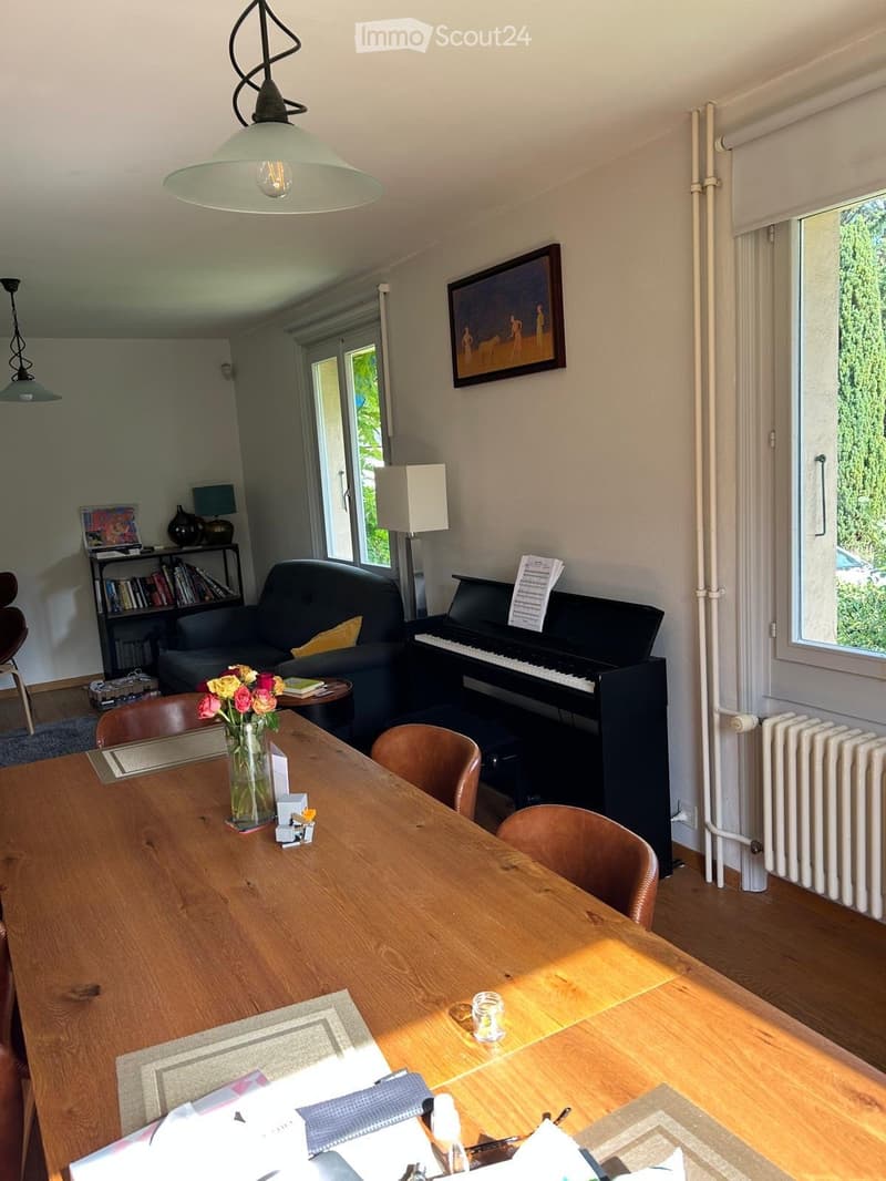 Bright living room with wooden floor, large windows, sofa, piano, dining table with chairs, bookshelf, and decorative items.