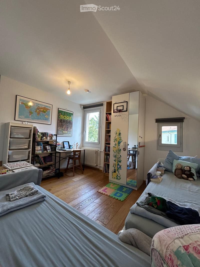 Cozy bedroom with a double bed, sofa, desk, chair, bookshelf, mirror, storage unit, laptop, and toys. The room features wooden flooring, large windows, and a slanted ceiling typical of an attic space.