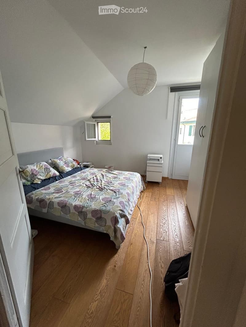 Cozy attic bedroom with wooden flooring, slanted ceiling, and large window. Features a double bed with colorful bedding, a nightstand, and a hanging paper lantern light fixture.