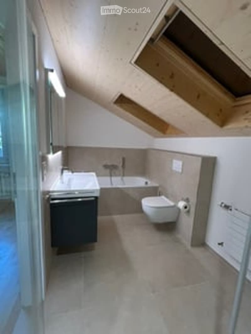 Modern bathroom with a sink, toilet, bathtub, and radiator. The room features exposed wooden beams on the ceiling and large windows for natural light.