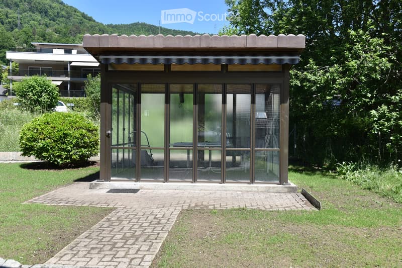 A small, modern outdoor structure with large glass panels and a striped awning, situated on a paved pathway surrounded by greenery and a grassy area.