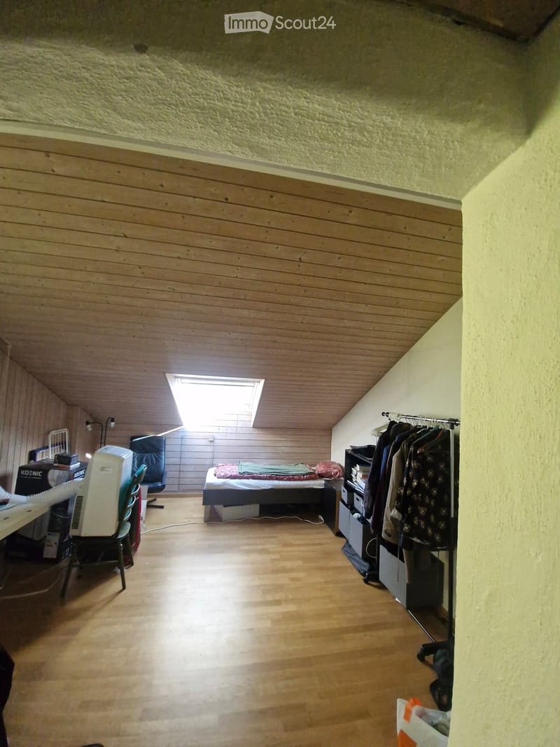 A furnished bedroom with wooden flooring, a bed, a desk, a chair, a wardrobe, and an air conditioner. The room has a slanted wooden ceiling and a skylight window.