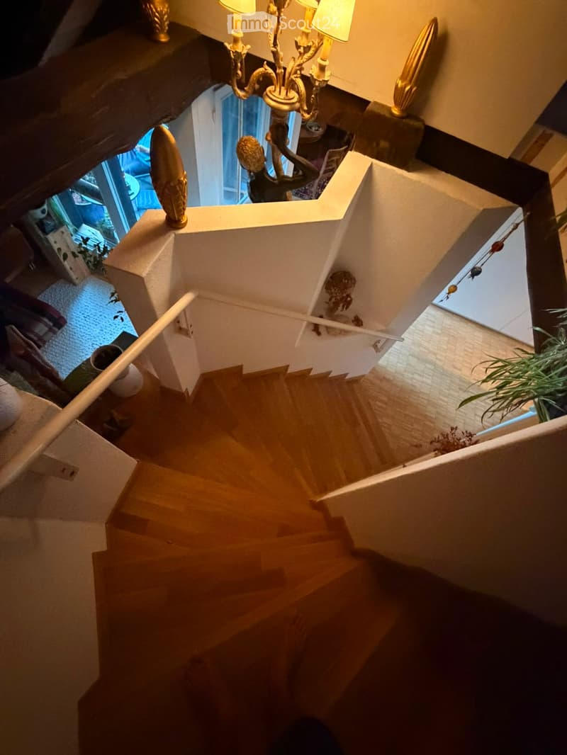 Wooden staircase with white railing, ornate chandelier, and decorative wall elements. Views of adjacent rooms and plants visible from the landing.