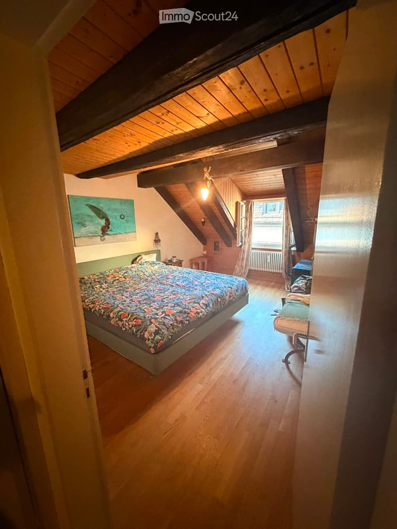 Cozy attic bedroom with wooden slanted ceilings, a double bed with floral bedding, a nightstand, a painting on the wall, and a small seating area with a chair and a small table.