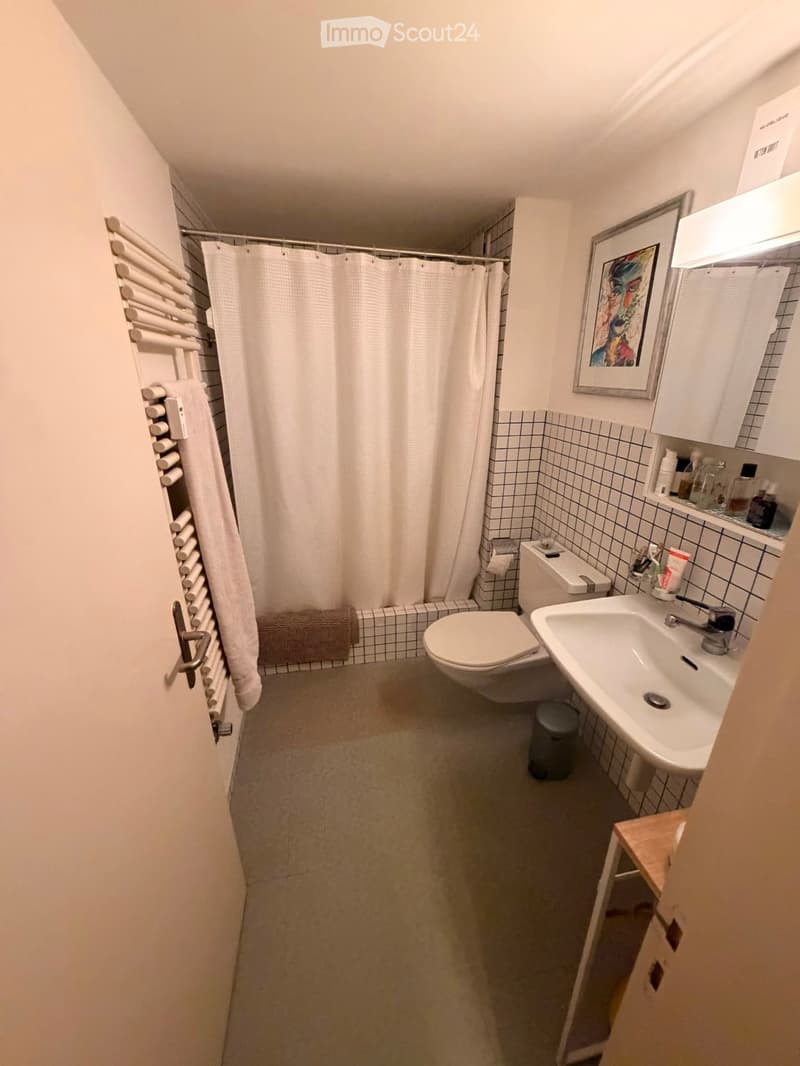 Modern bathroom with white tiles, a toilet, sink, mirror, and shower curtain. Includes a towel rack, picture frame, and small cabinet.