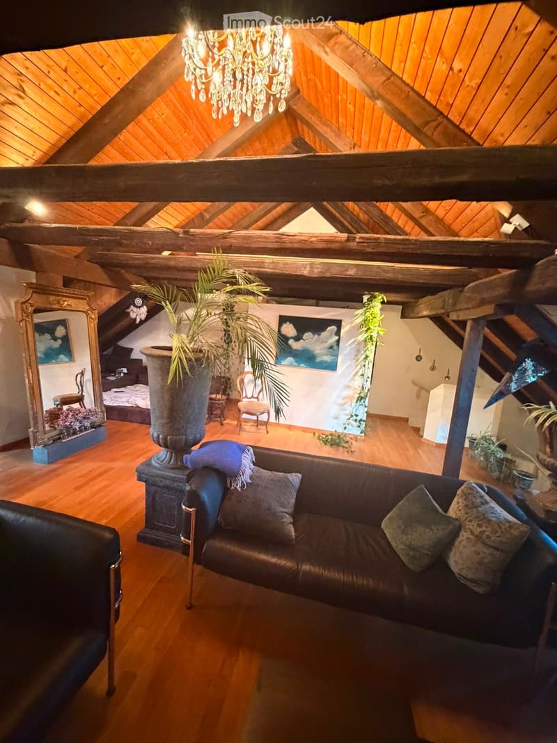 Loft-style living room with wooden beams, exposed ceiling, chandelier, leather sofa, wooden chairs, large mirror, and decorative plants.