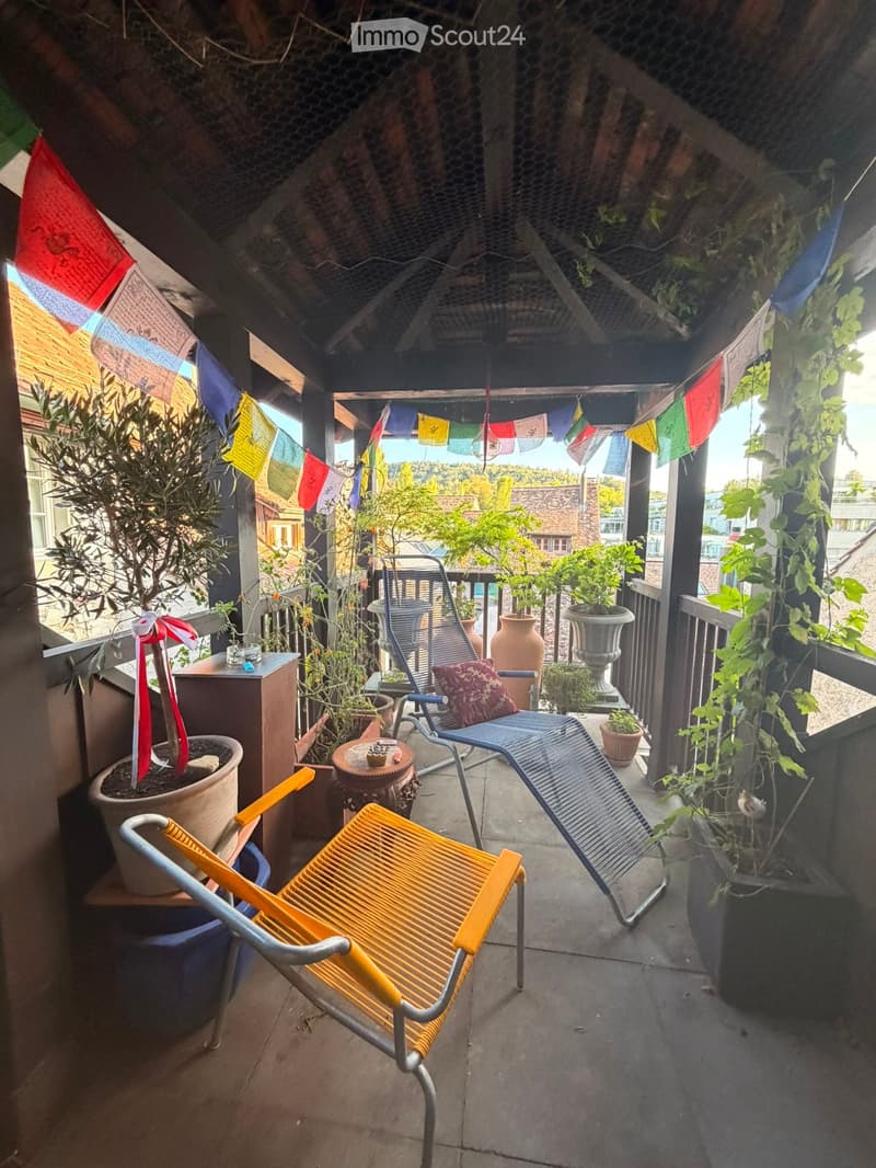 Balcony with colorful hanging flags, potted plants, and two chairs.