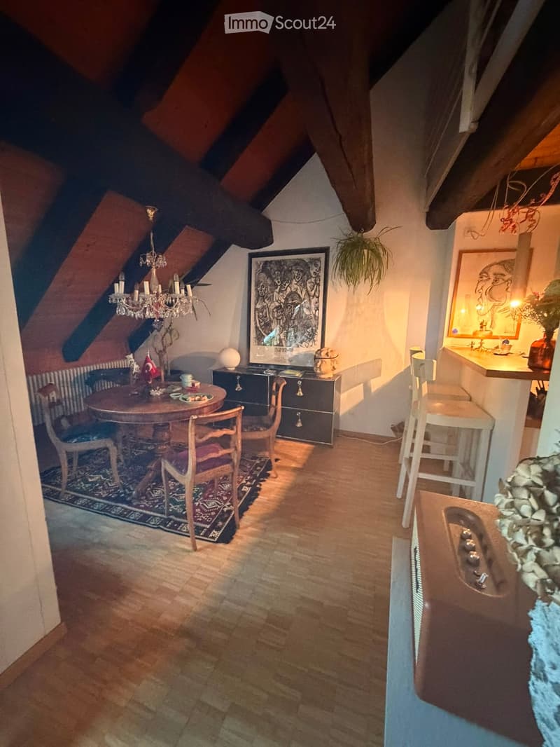 Attic room with wooden floors, slanted ceilings, a dining area with a round table and chairs, a chandelier, a cabinet, and a small kitchen area with a sink and stools.