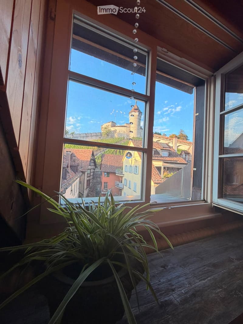 Balcony with wooden floor, large windows offering a view of a historic town with a tower and traditional houses.
