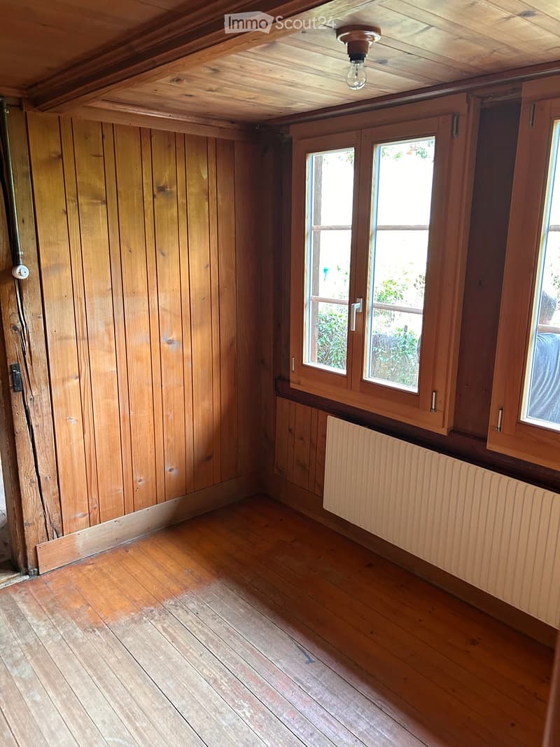 Empty room with wooden walls, wooden floor, and two windows with wooden frames. There is a radiator under one window and a light fixture on the ceiling.