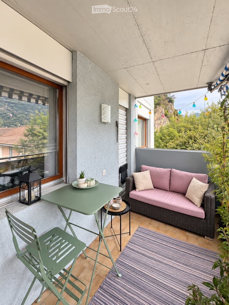 Balcony with a pink sofa, green table and chairs, small rug, lantern, and potted plant, overlooking a scenic mountain view.