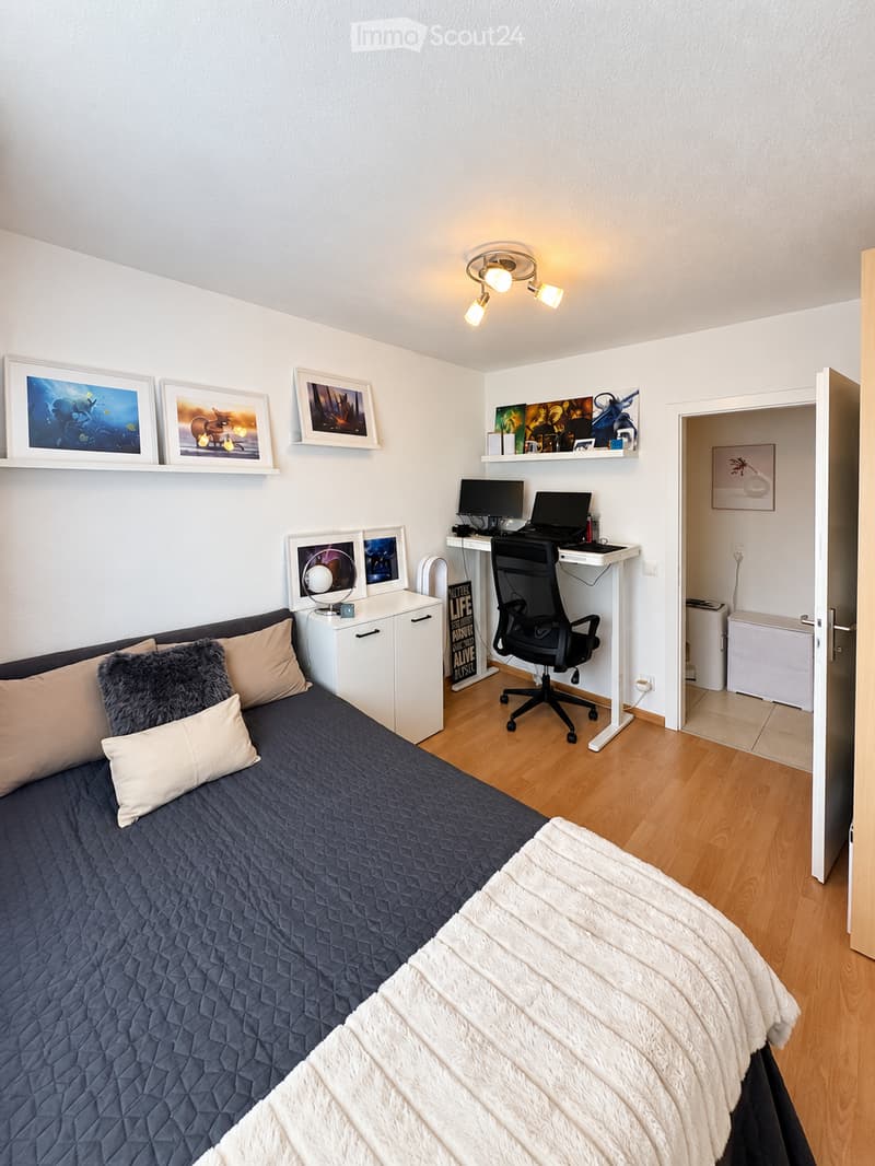 A modern, well-lit bedroom with a single bed, a desk with a laptop and computer monitor, a chair, a small cabinet, and several framed pictures on the wall. The room features light wooden flooring and white walls.