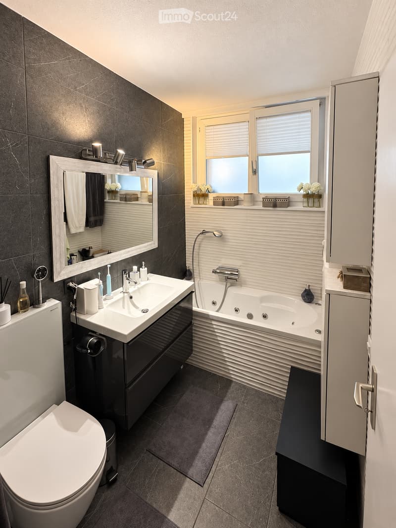 Modern bathroom with a double sink, bathtub, toilet, large mirror, and window with blinds. Includes decorative vases and a light fixture above the mirror.