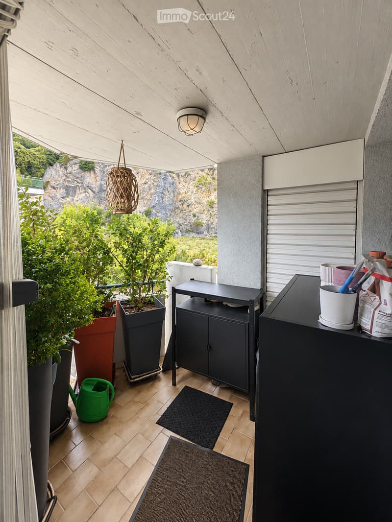 Balcony with wooden flooring, black table, potted plants, and a view of greenery and rocky terrain.