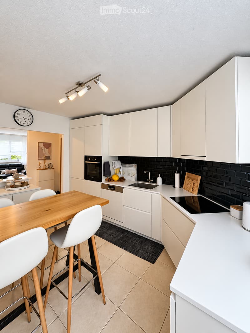 Modern kitchen with white cabinets, a wooden dining table, white chairs, and a black brick backsplash. Features include an oven, sink, and countertop items.