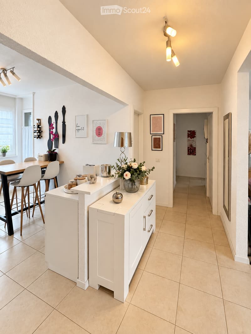 Modern dining area with a white cabinet, vase with flowers, lamp, framed pictures, and a dining table with chairs.