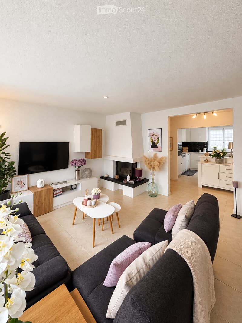 Modern living room with a dark grey sectional sofa, white coffee table, fireplace, flat-screen TV, and decorative plants.