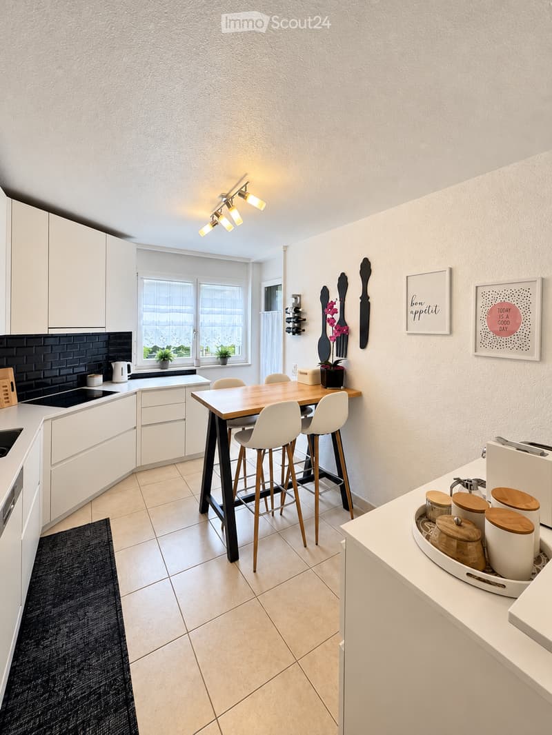 Modern kitchen with white cabinets, a wooden dining table with four white stools, a black tiled backsplash, and a kitchen island with a sink and storage. The space features a tiled floor, framed artwork, and a small potted plant on the windowsill.