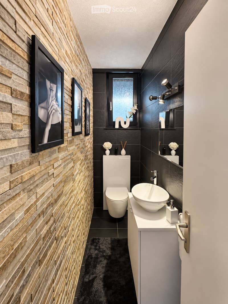 Modern bathroom with a white toilet, a white sink with a modern faucet, a large mirror, and a framed picture on the wall. The walls are tiled in a mix of light wood and dark tiles, and the floor is black with a plush black rug.