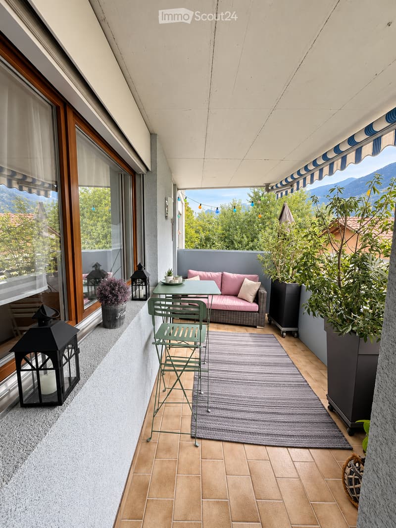 Balcony with tiled floor, sofa, small table, chairs, rug, and decorative lanterns, overlooking a scenic mountain view.