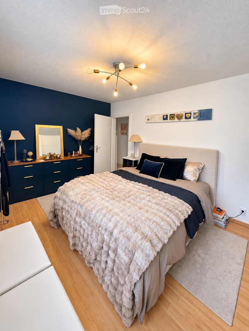 Modern bedroom with a large bed, dark blue walls, a fur throw, and a stylish dresser with a mirror. The room features a chandelier light fixture and a small side table with a lamp.