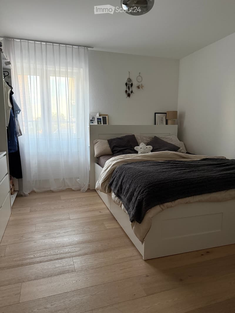 Modern bedroom with a large bed, pillows, blanket, nightstand with lamp, picture frames, and a curtained window. Wooden flooring and white walls.